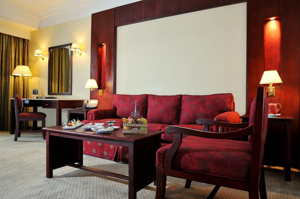 Safir Hotel Cairo Giza Стая снимка The photo shows a well-decorated hotel room or sitting area. In the center, there is a red couch with matching armchairs around it. A wooden coffee table is placed in front of the couch, and it holds some decorative items and snacks. The walls are ad