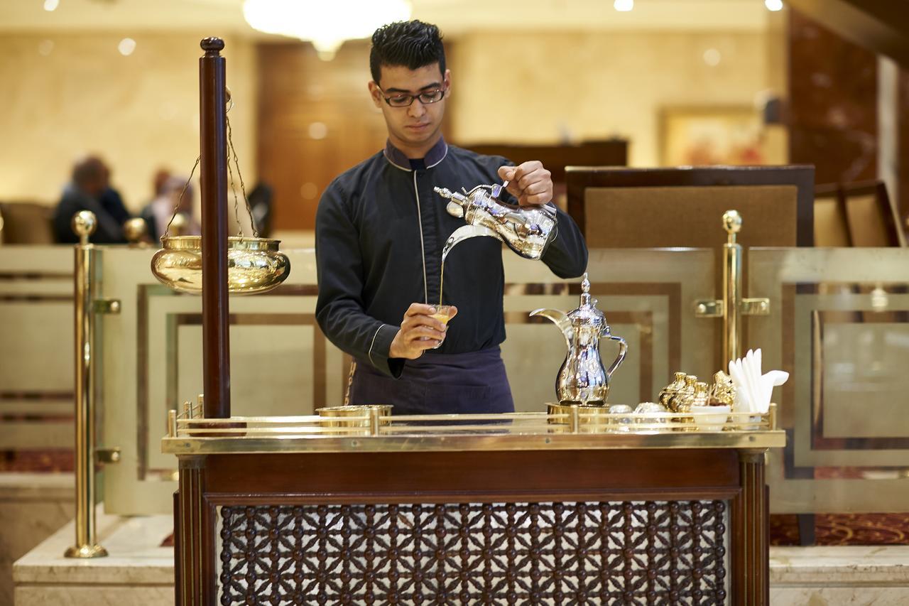 Safir Hotel Cairo Giza Екстериор снимка The photo depicts a person at a service counter, pouring tea from a traditional teapot. The individual appears to be dressed in a formal, dark outfit, and is focused on the task of pouring. The counter features ornate decorations and various tea-serv