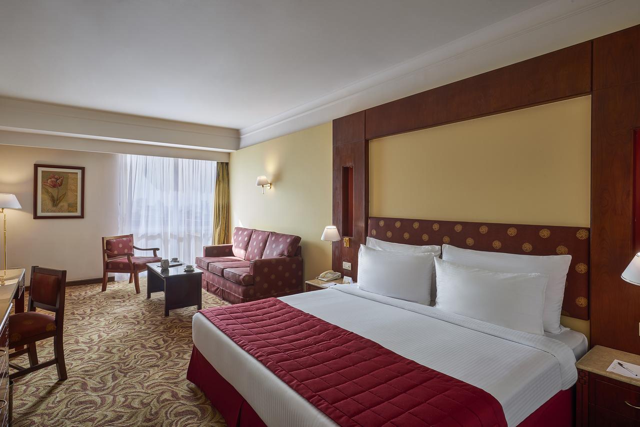 Safir Hotel Cairo Giza Екстериор снимка The photo shows a hotel room featuring a spacious layout. In the center, there is a large bed with white bedding and a red decorative coverlet. To one side, there is a seating area that includes a sofa and two armchairs, all set around a small coffee