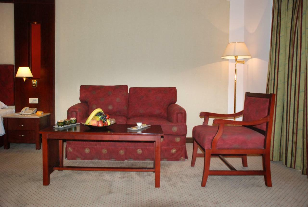 Safir Hotel Cairo Giza Екстериор снимка The photo shows a cozy seating area in a hotel room. There is a red patterned sofa and a matching armchair placed around a wooden coffee table. On the table, there are some snacks or fruits displayed. A floor lamp is positioned beside the sofa, and t