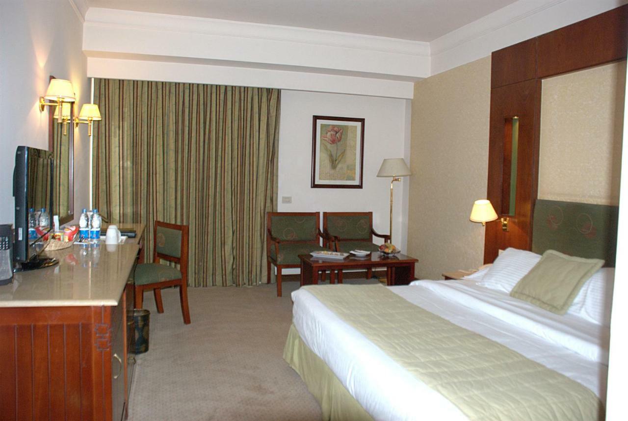 Safir Hotel Cairo Giza Стая снимка The photo shows a hotel room. It features a bed with white linens and a green throw. On one side of the bed, there's a small bedside lamp. Opposite the bed is a desk with a mirror above it, alongside a couple of chairs and a small table. The room has