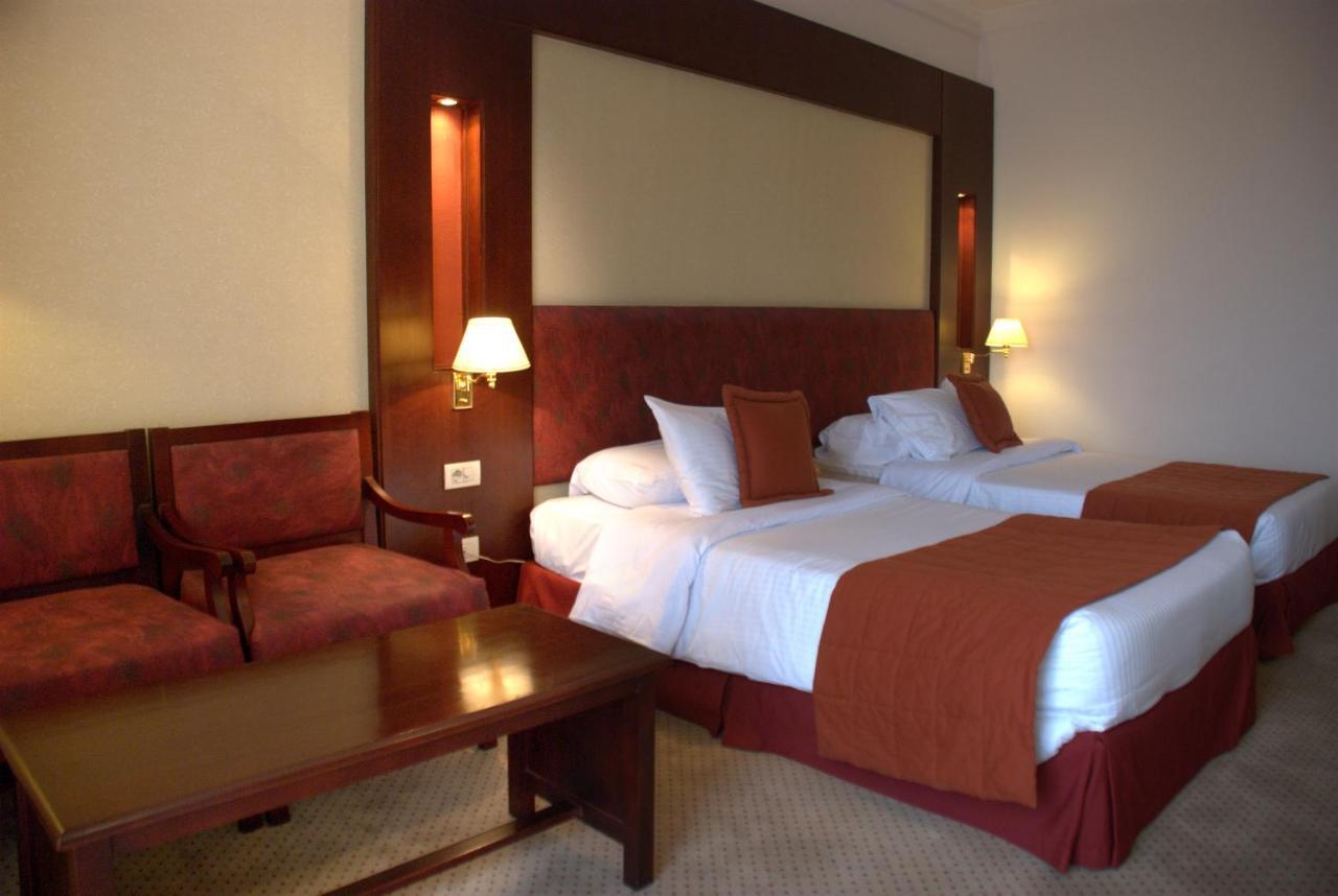 Safir Hotel Cairo Giza Екстериор снимка The photo shows a hotel room featuring two twin beds with white linens and orange bedspreads. There is a headboard behind the beds, along with wall-mounted lamps on either side. In the corner, there are two upholstered chairs and a wooden coffee tabl