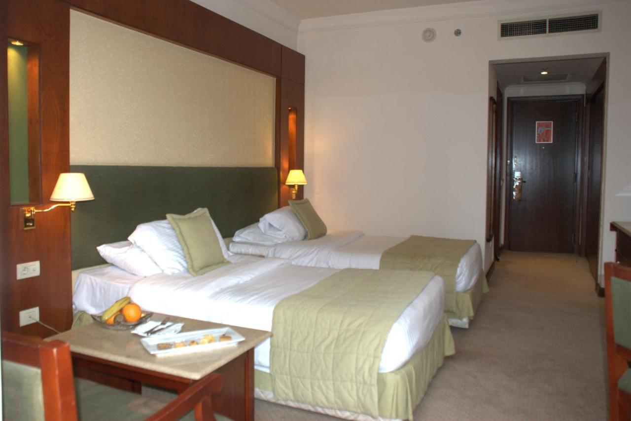 Safir Hotel Cairo Giza Стая снимка The photo shows a hotel room featuring two single beds with light green bedspreads and white linens. There are bedside lamps on either side of the beds, and the headboard has a soft, textured design. A small seating area is visible with a wooden chai